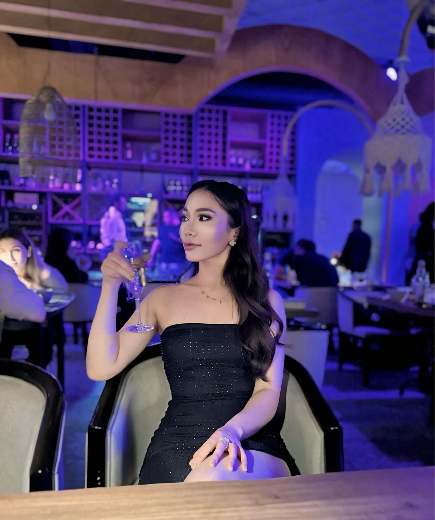 A woman sits at a lounge table under purple lighting, wearing a strapless black mini dress and holding a stemmed glass.