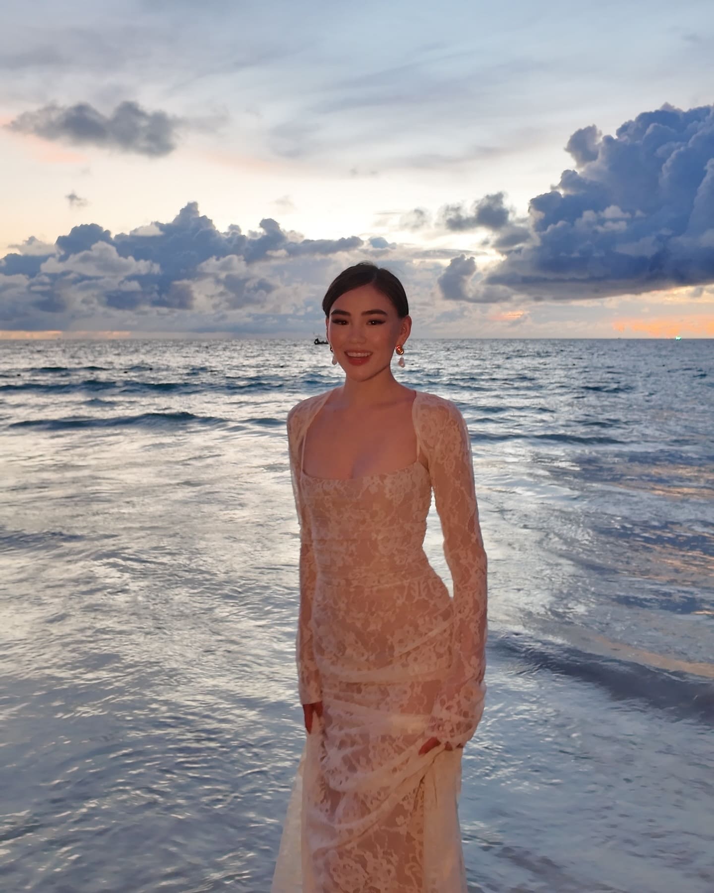 A woman stands in shallow ocean water at sunset, wearing a long-sleeved, sheer white lace gown with a square neckline.