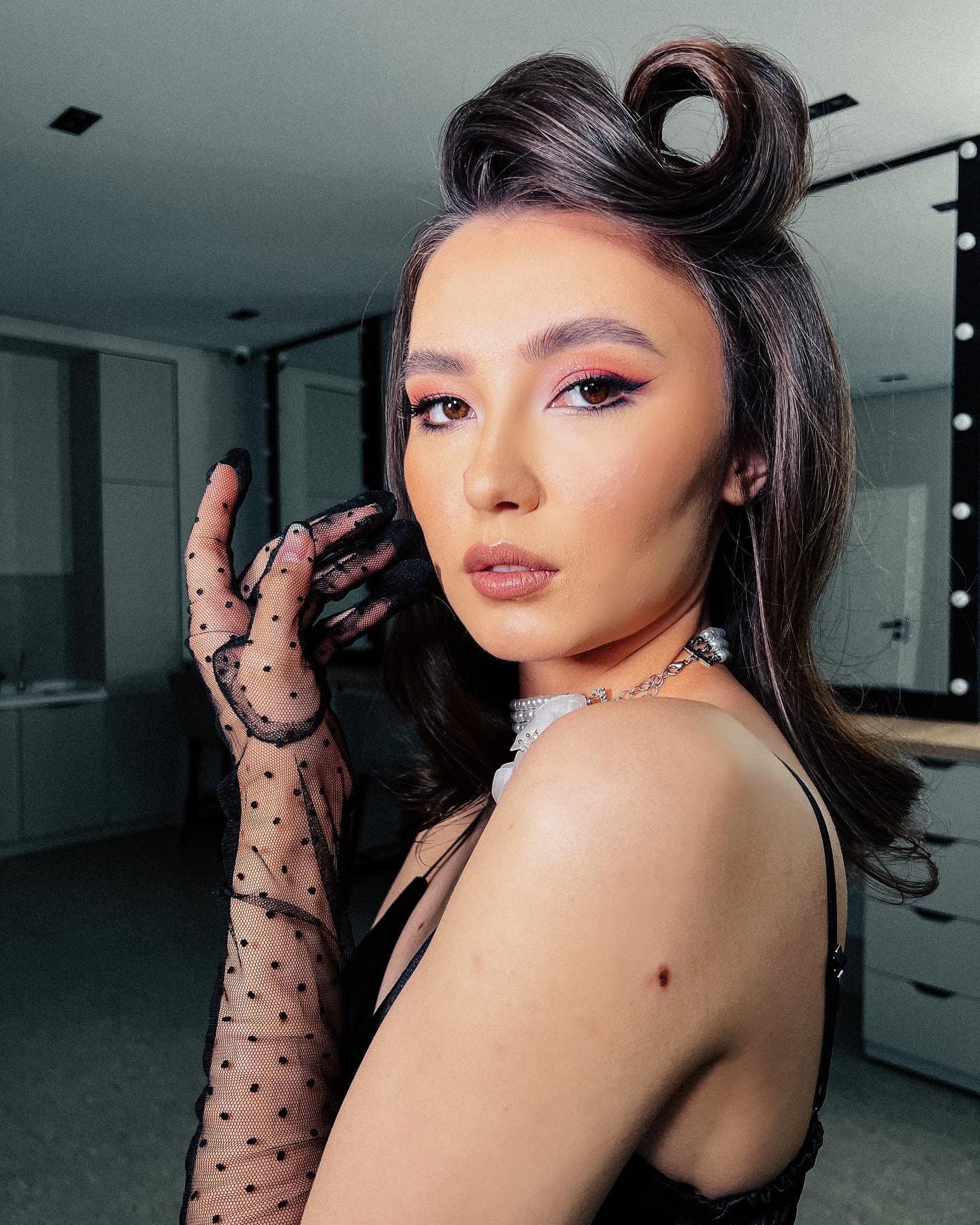 女子梳复古波浪盘发，穿黑色细带上衣，戴白色颈圈及黑色波点透明长手套，在更衣室内摆姿势。