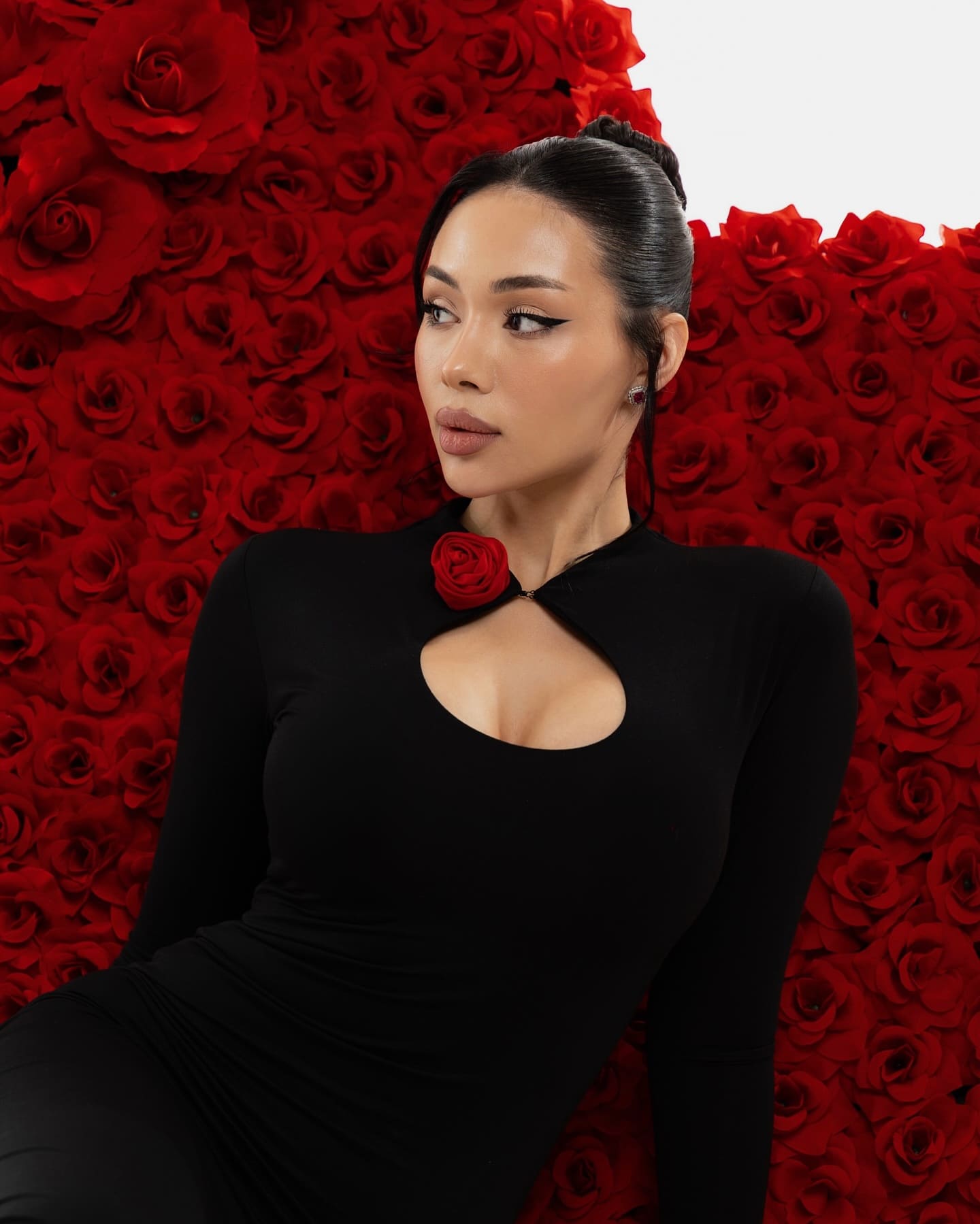 Dilyara wears a black dress with a red rose cutout, posing against a wall of red roses.