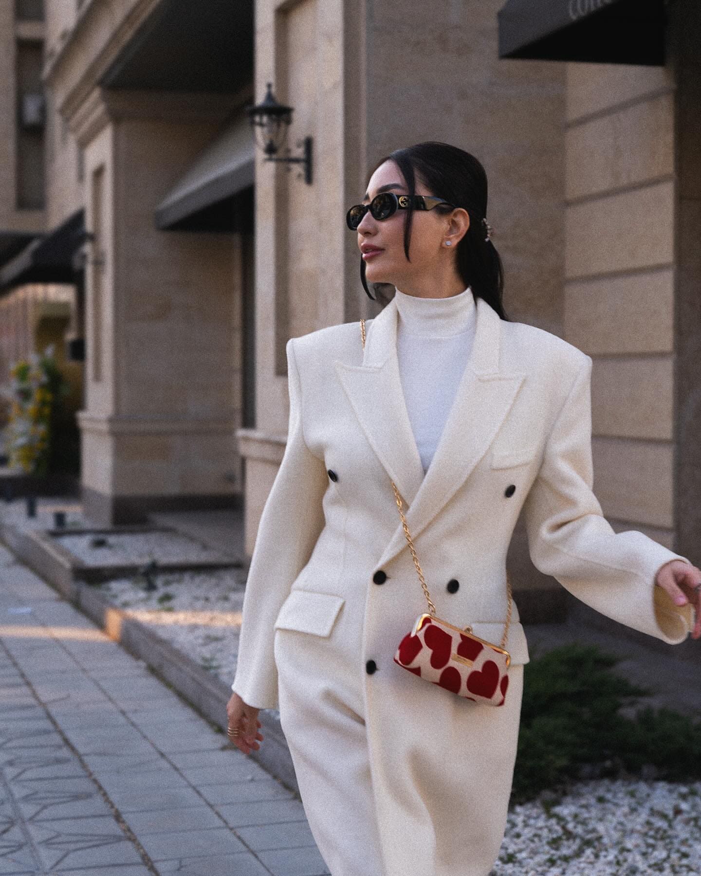 A woman in a cream coat and sunglasses walks a city sidewalk carrying a heart-patterned bag.