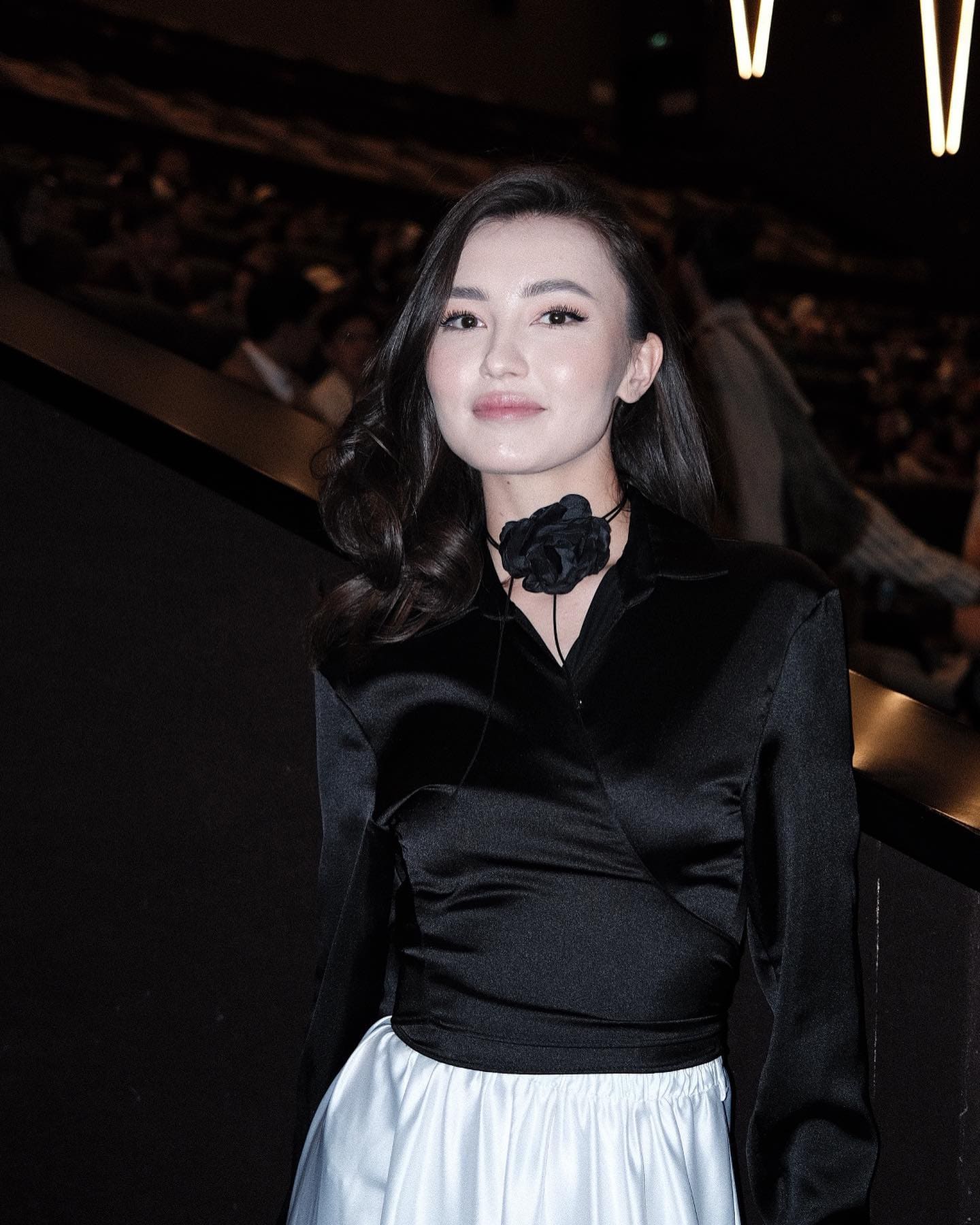 长卷发女子正面站立，身着黑色缎面长袖衬衫配白色裙子，佩戴黑色大花颈圈，背景昏暗带暖光。