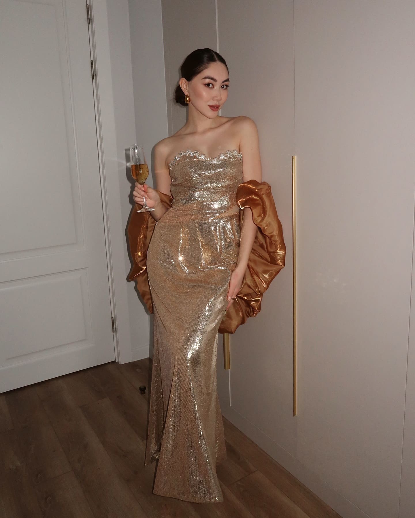 Woman in a gold sequined gown and satin shawl stands indoors, holding a glass of champagne.