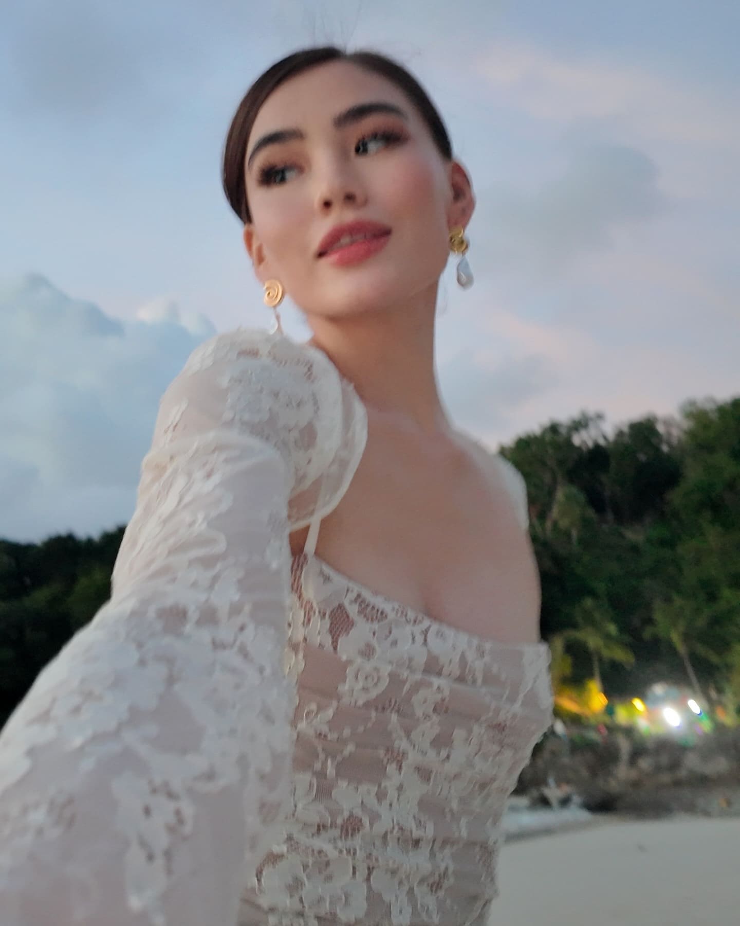 A woman poses outdoors against trees and a cloudy sky, wearing a white lace off-the-shoulder dress with long sleeves and gold drop earrings.