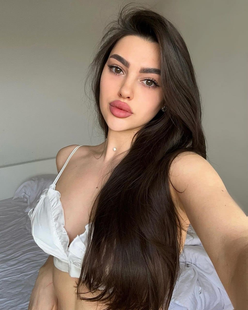 Amalia sits on a bed with white sheets, wearing a white bralette, taking a selfie against a plain wall.