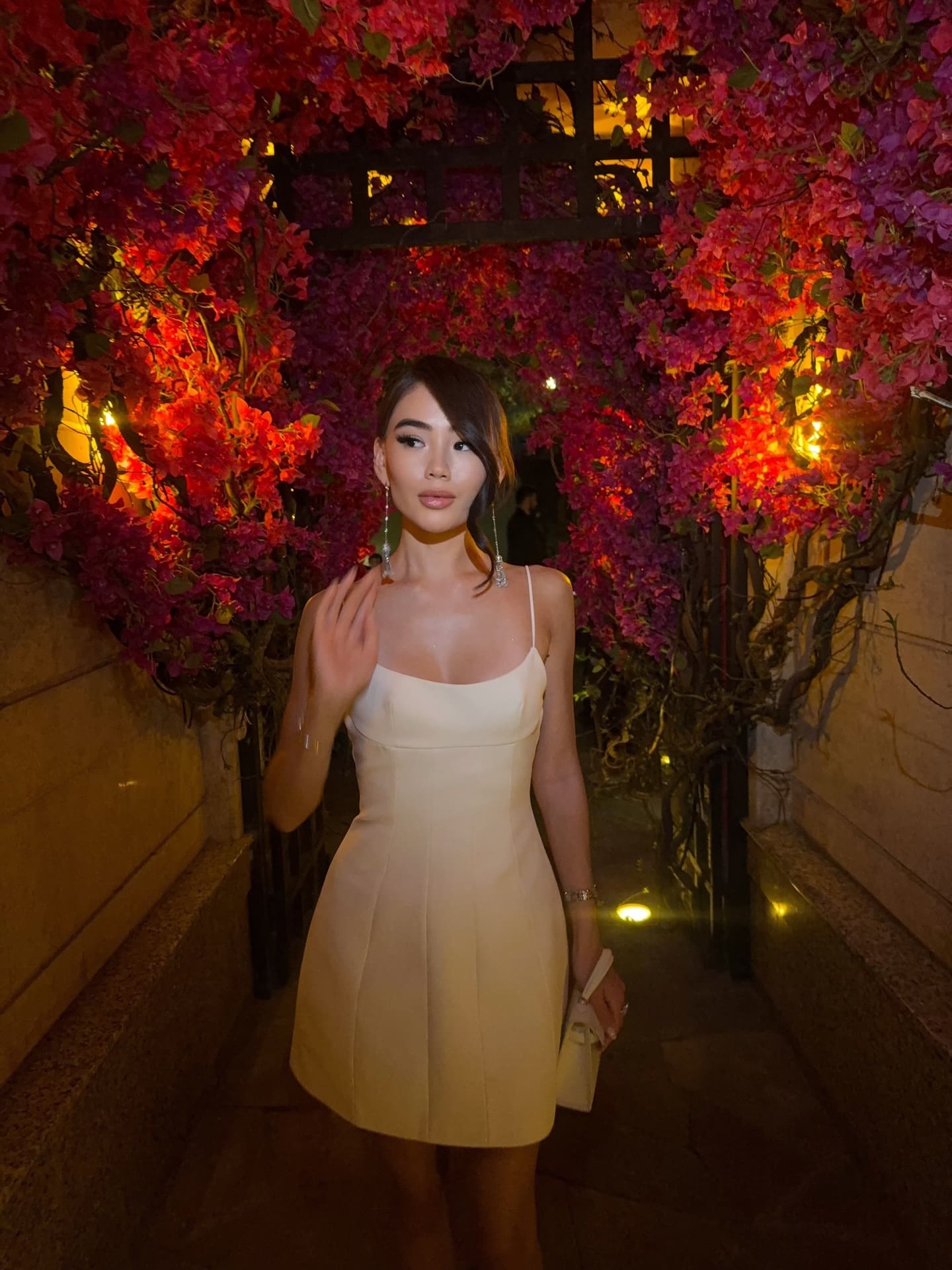 A woman stands in a pathway framed by pink and red bougainvillea, wearing a cream mini dress with spaghetti straps and holding a small clutch.
