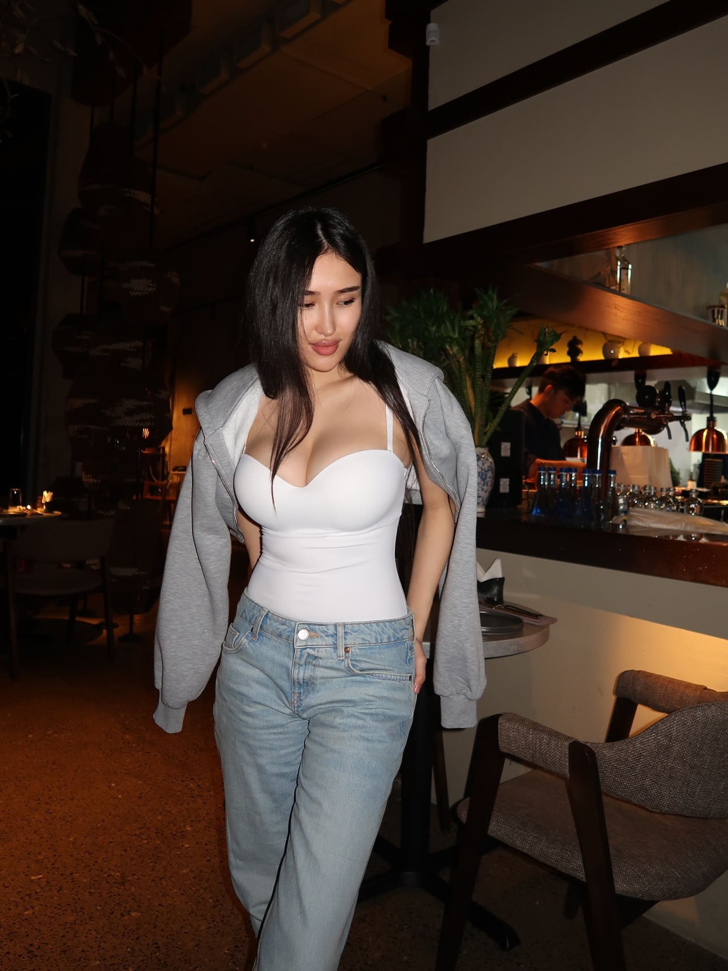 A woman stands indoors wearing a white bodysuit, light-wash jeans, and a grey hoodie draped loosely off her shoulders.