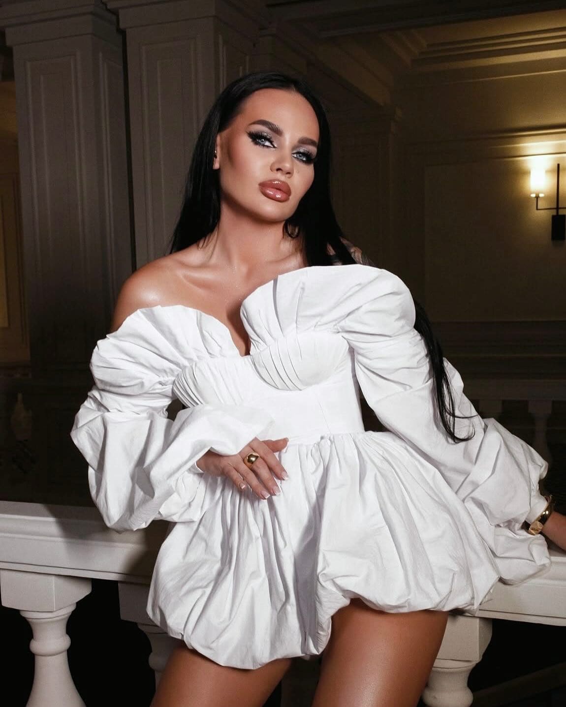Arina leans against a white balustrade indoors, wearing a white off-the-shoulder mini dress with voluminous sleeves.
