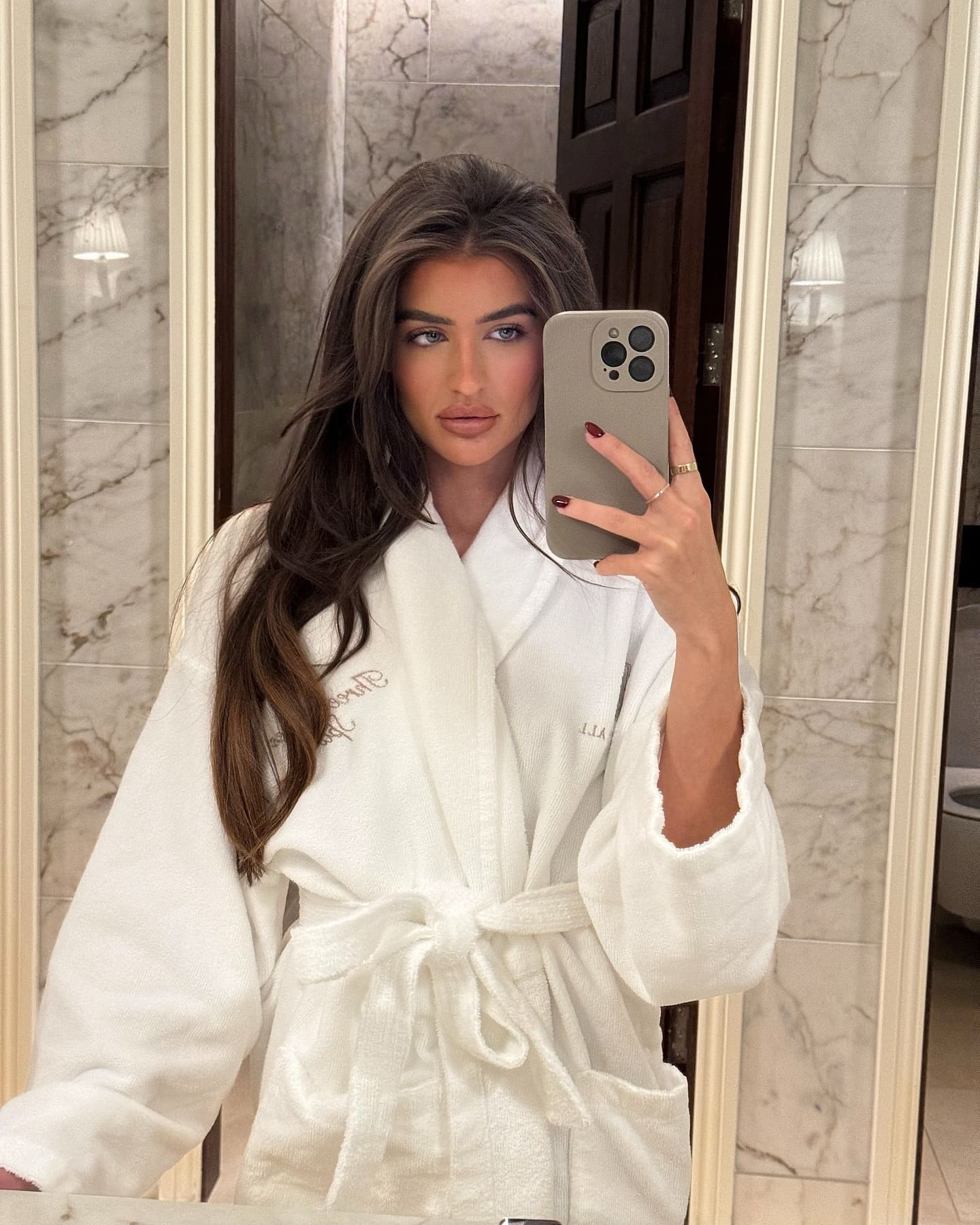 Alisa stands in a marble-tiled bathroom, taking a mirror selfie with a gray smartphone while wearing a white waffle-knit bathrobe.