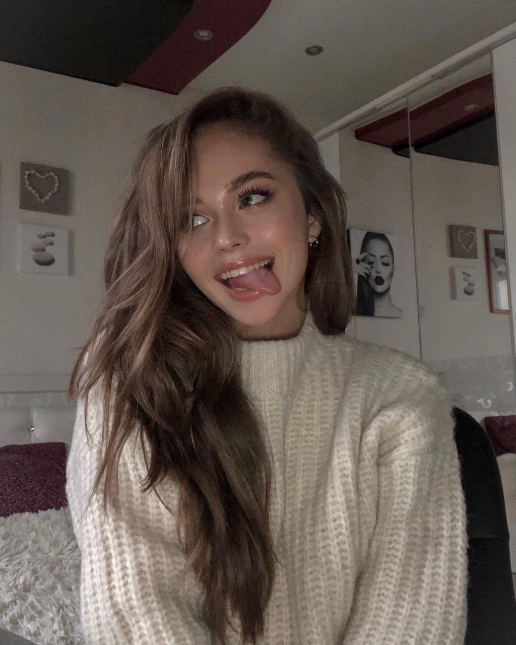 Woman in a chunky cream knit sweater sticks her tongue out while looking sideways in a bedroom.
