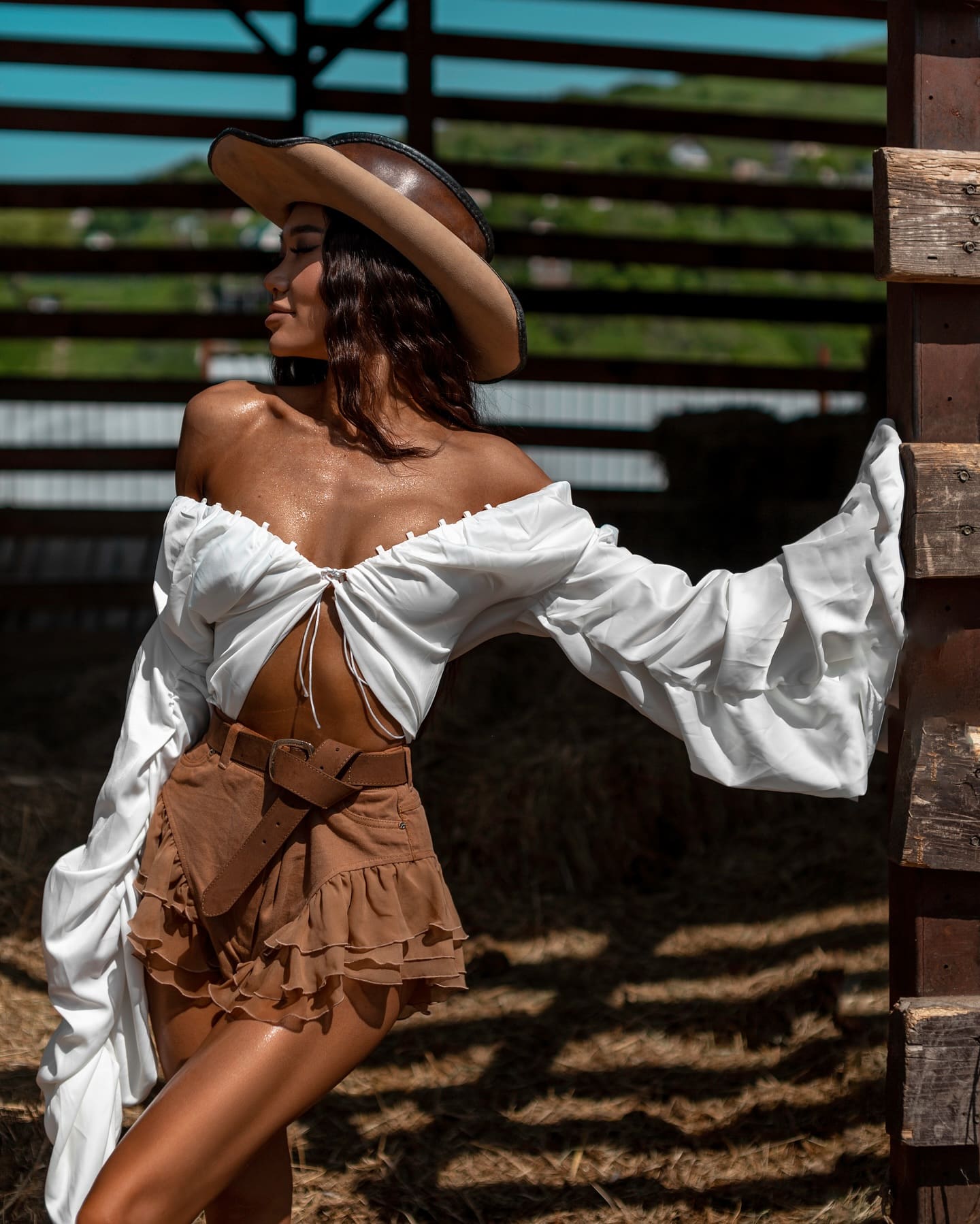 Posing in profile by a barn, she wears a brown cowboy hat, white off-shoulder blouse, and tiered brown mini skirt.