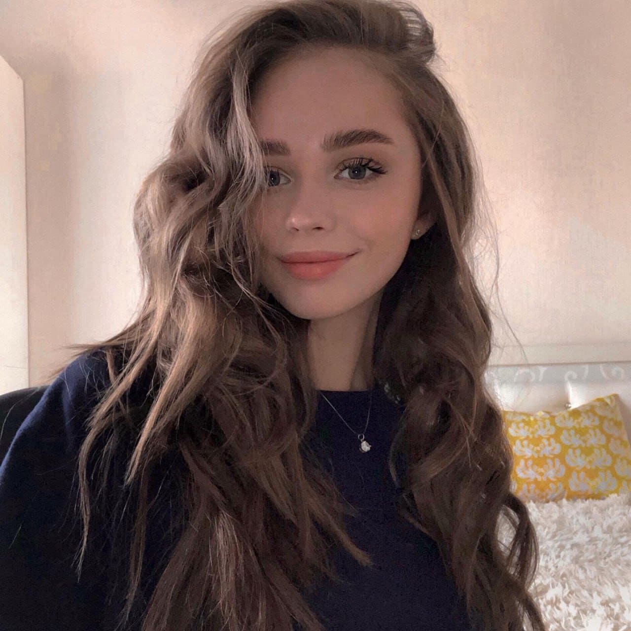 Woman with brown hair in a navy top and necklace poses indoors against a light wall with a yellow pillow.