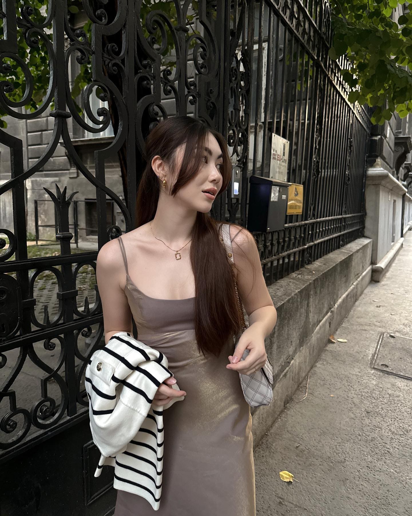 Woman in a taupe satin slip dress stands on a sidewalk by an iron fence, holding a striped garment.