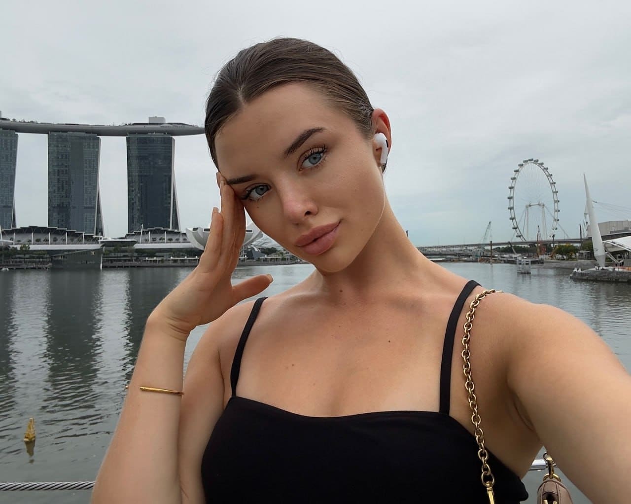 Emmeline poses with a hand to her temple in a black top, set against a waterfront with a hotel and Ferris wheel.