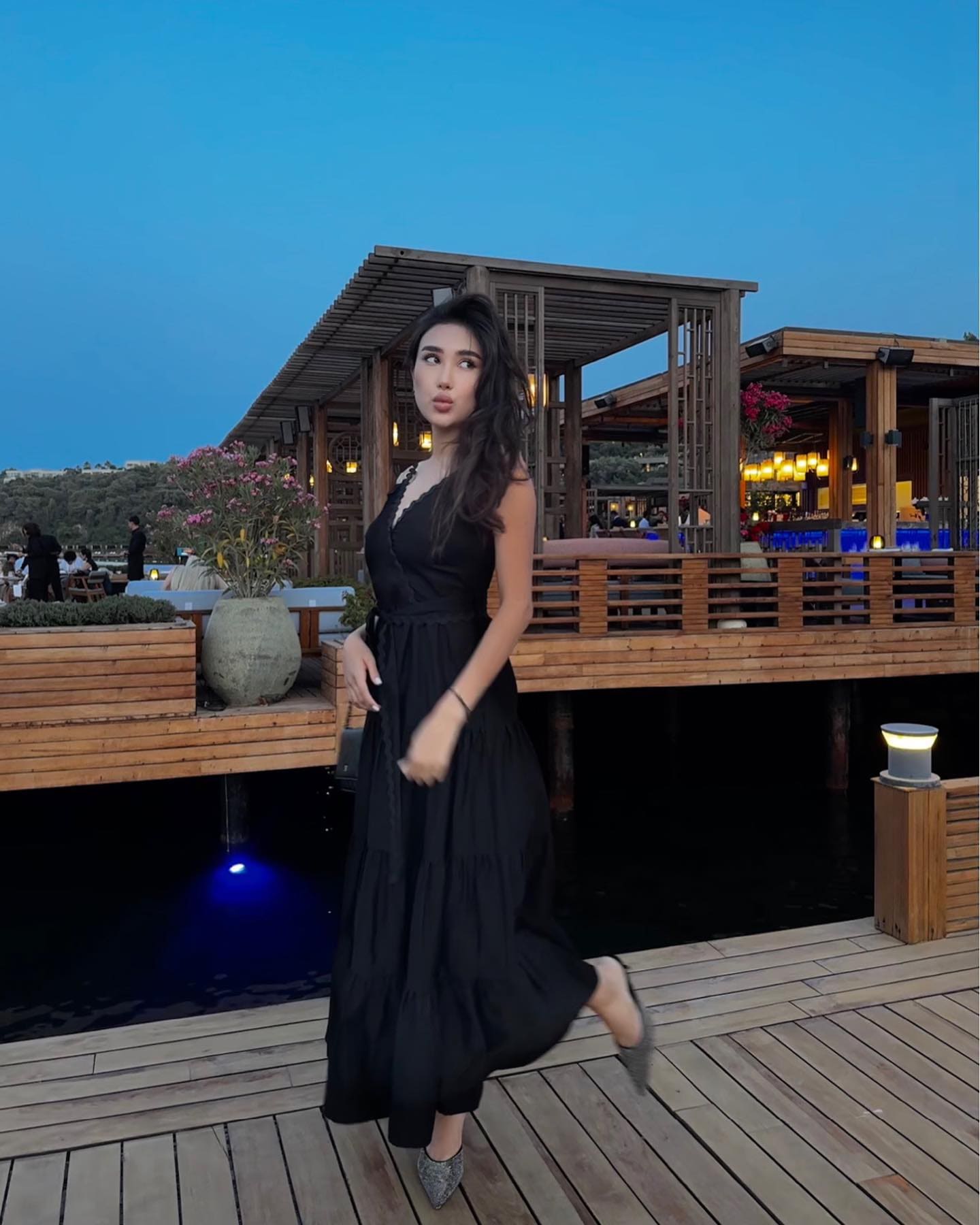 A woman in a black sleeveless dress with lace trim stands on a boardwalk at dusk, posing with one leg raised near a lit waterfront structure.