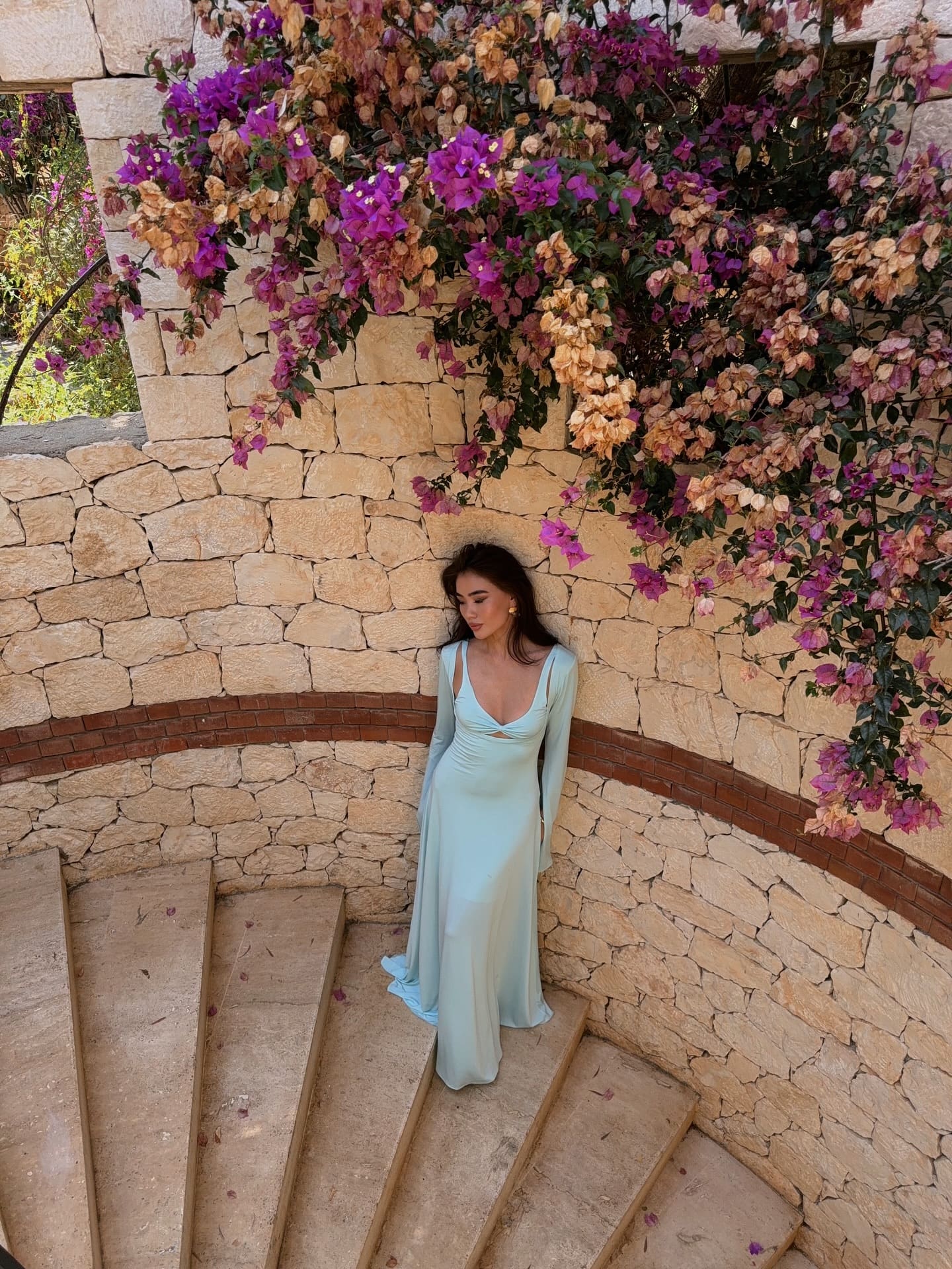 A woman stands on stone steps beneath purple bougainvillea, wearing a floor-length light blue gown with long sleeves and a deep V-neckline.