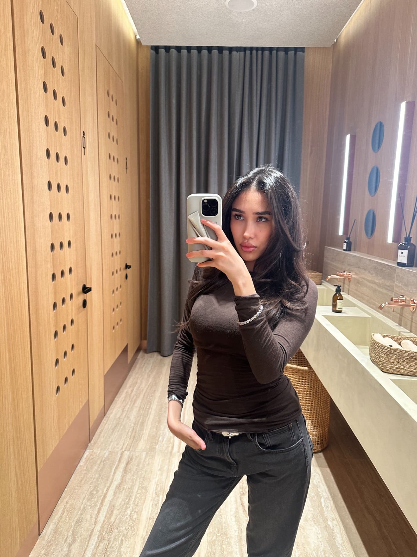 Aknur takes a mirror selfie in a wood-paneled restroom, wearing a brown long-sleeved top and dark grey jeans with one hand in her pocket.