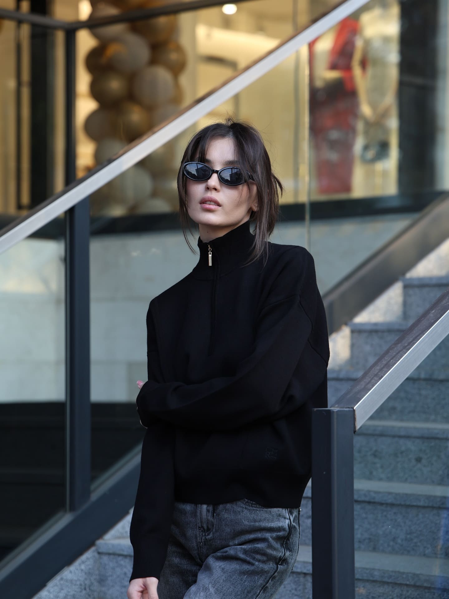 女子戴黑色椭圆墨镜，穿黑色长袖半拉链上衣与灰色牛仔裤，双臂交叉站在户外混凝土台阶上。