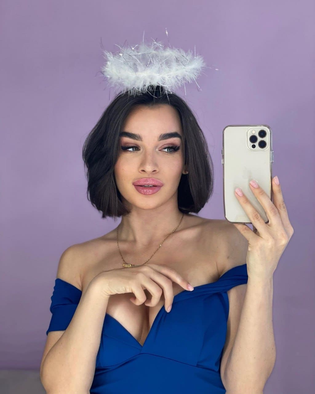 Estelle takes a mirror selfie in a blue off-shoulder dress and feather halo against a purple background.