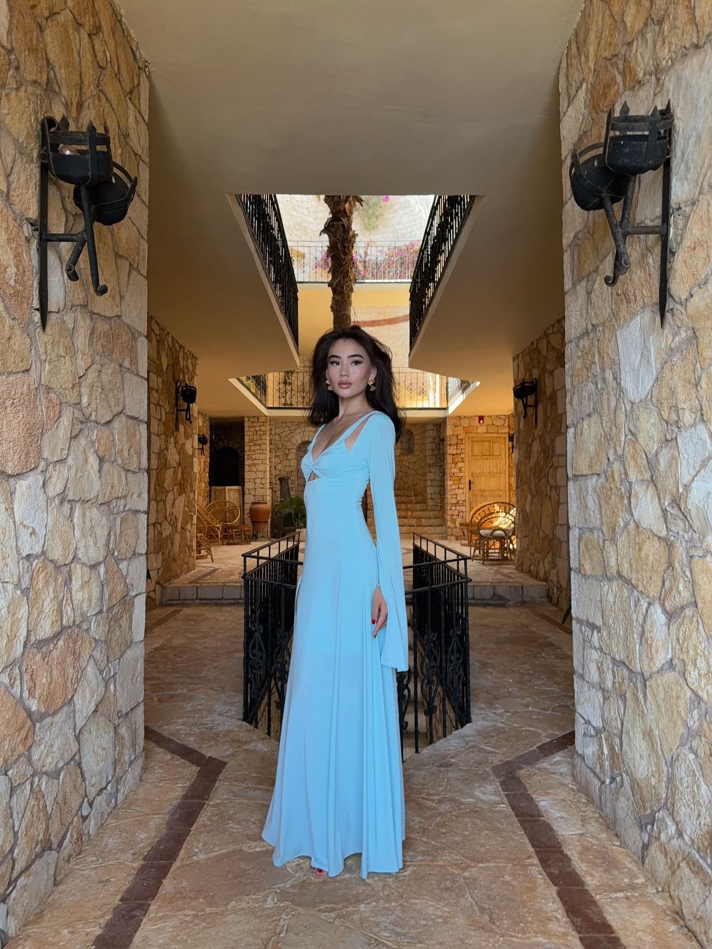 A woman with long dark hair stands in a stone corridor wearing a floor-length light blue gown with long sleeves, a deep V-neckline, and waist cutouts.
