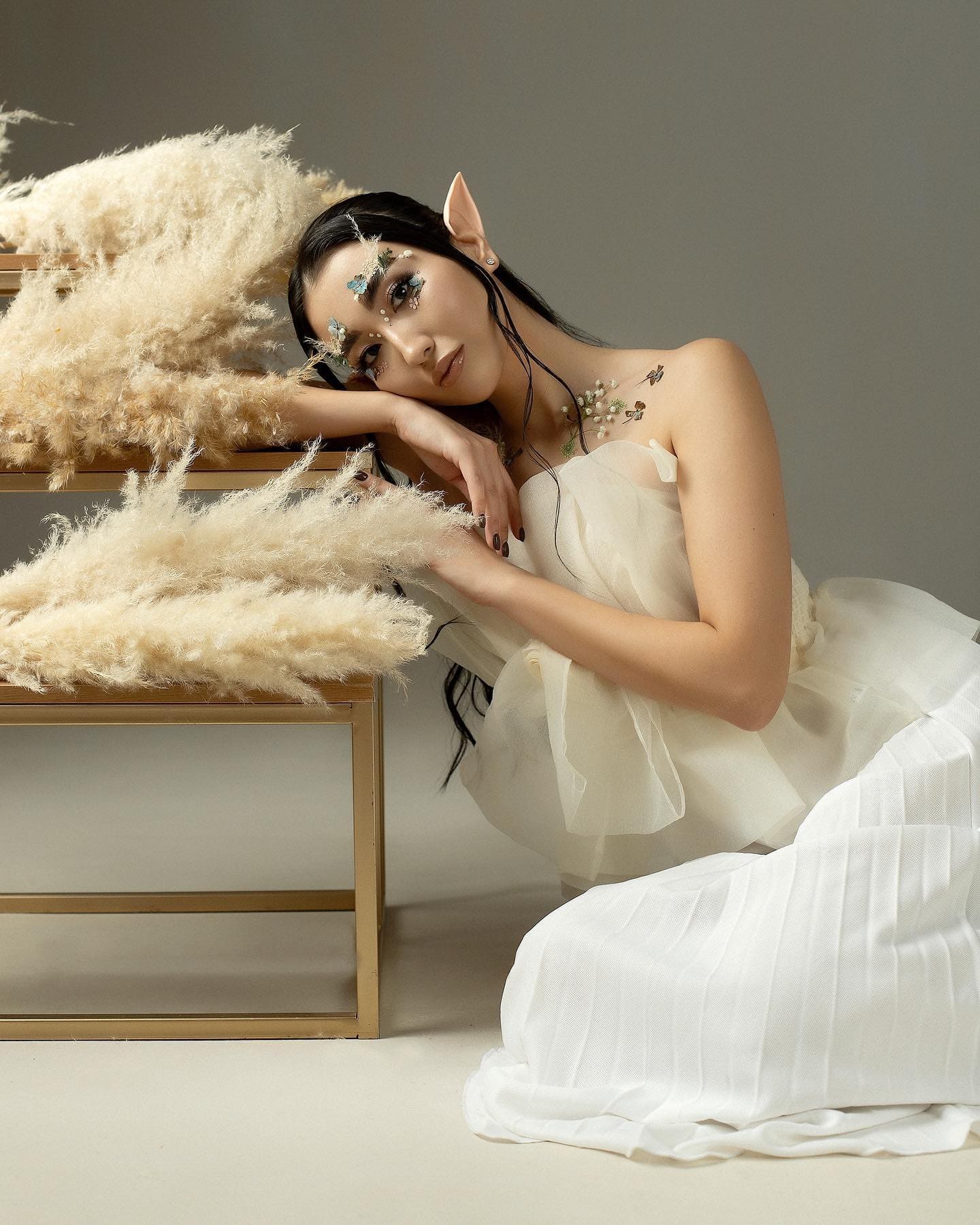 A woman with elf ears in a white tulle gown reclines on a gold bench draped with pampas grass.
