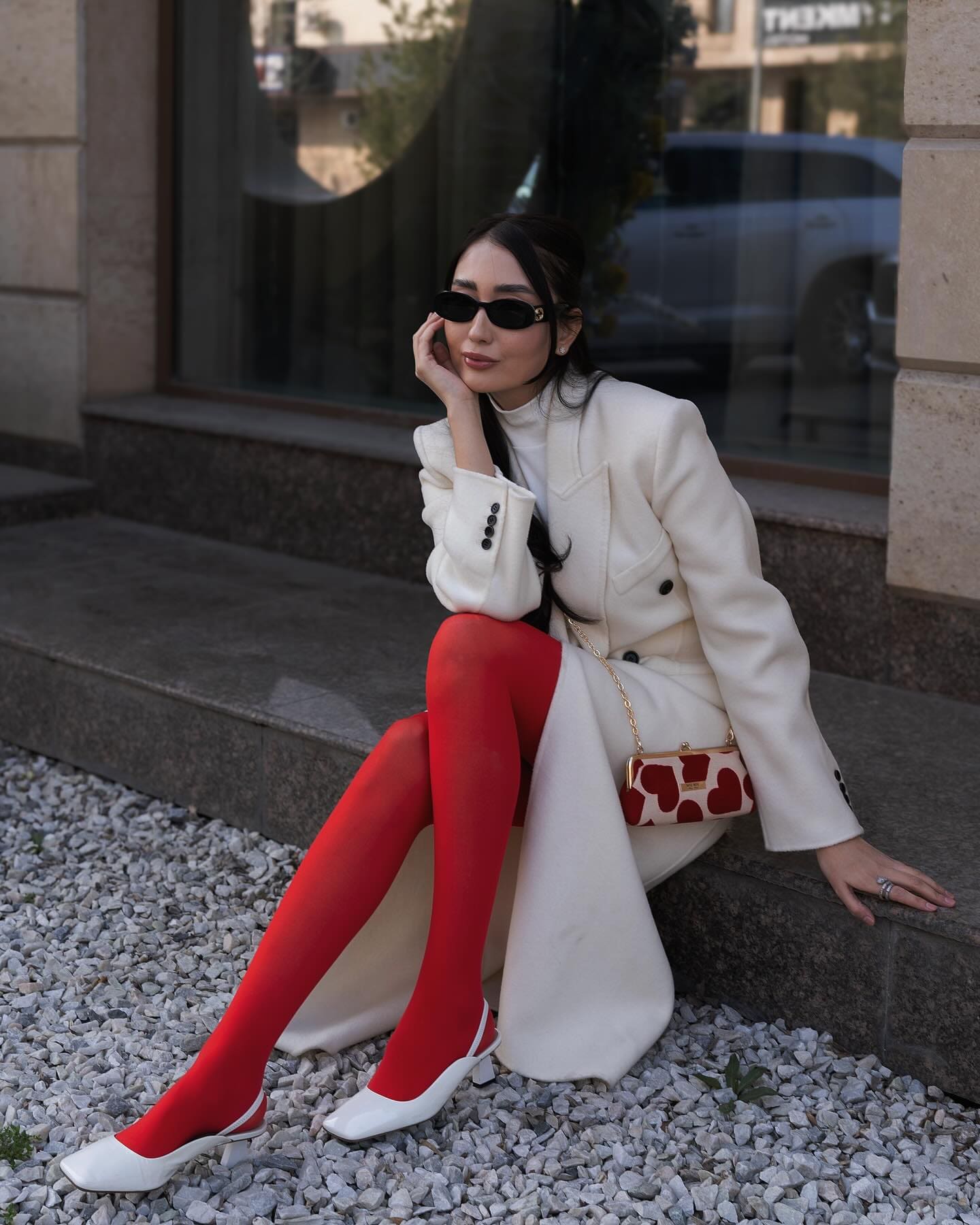 A woman in a cream coat and red tights sits on a stone ledge wearing sunglasses and white heels.