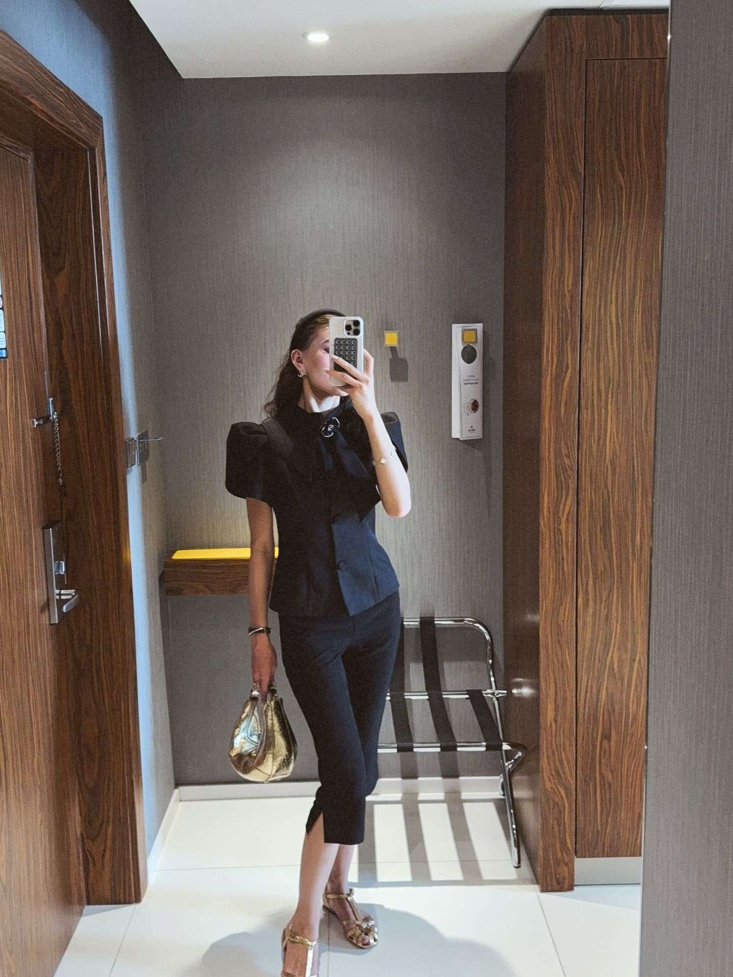 A woman takes a mirror selfie in a hallway, wearing a black cropped jacket over a white shirt, black trousers, gold heels, and holding a gold handbag.