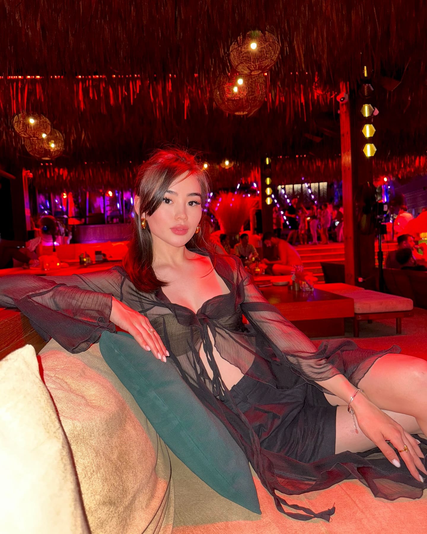 A woman sits on a lounge sofa in a dimly lit venue with red lighting, wearing a sheer black top over a bra and shorts, legs extended to the side.