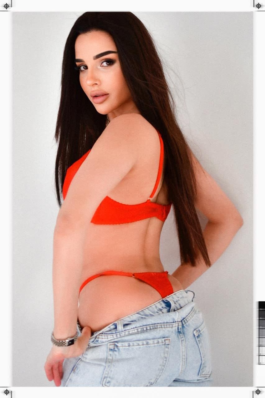 Profile view of a woman in red bikini and jeans pulled down to thighs against grey background.