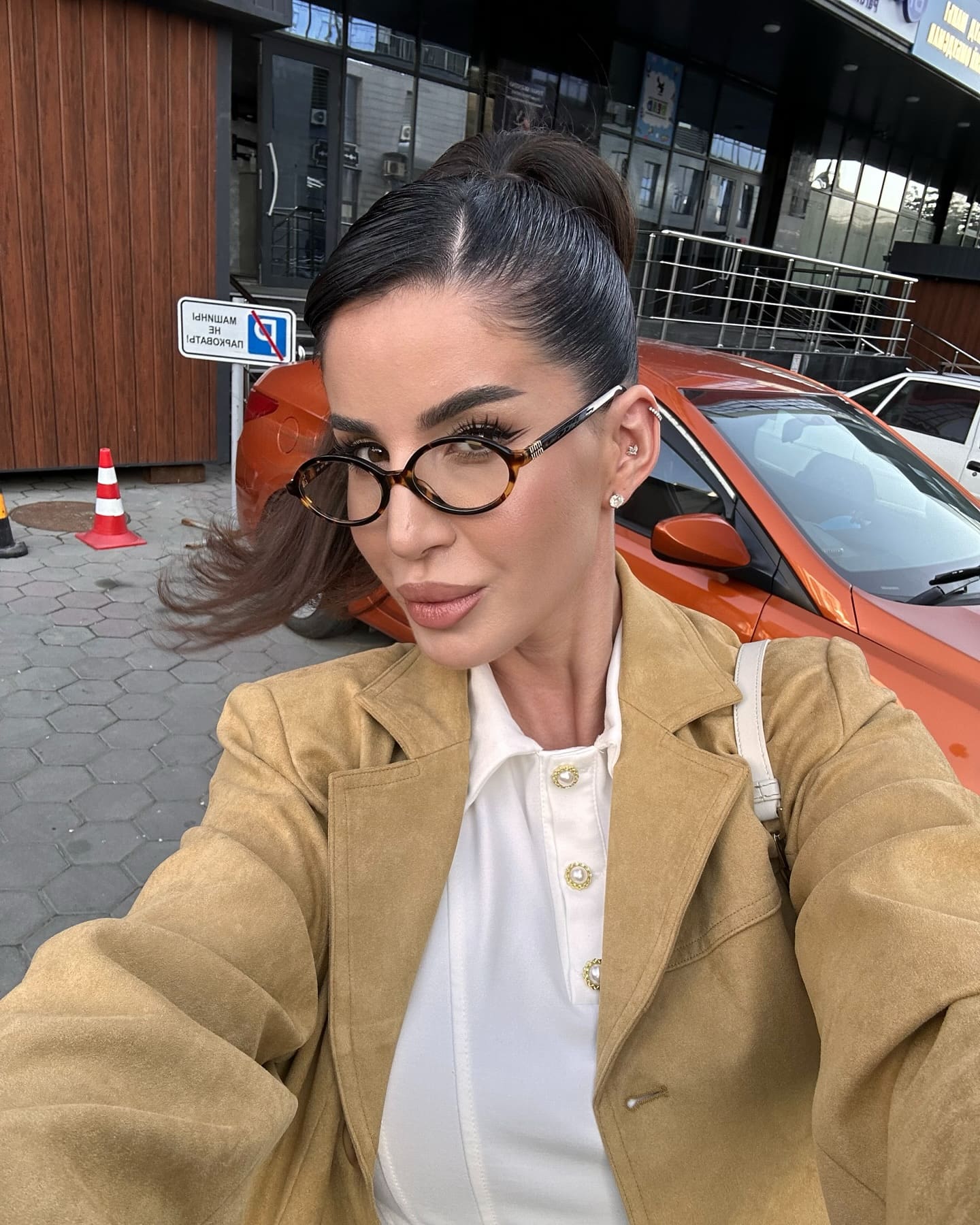 Dinara takes an outdoor selfie in a white shirt, tan suede jacket, and glasses, with an orange car behind her.