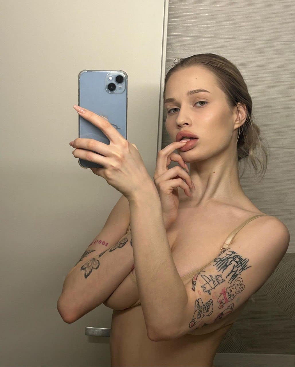 A woman with brown hair in a nude bra touches her lips while taking a mirror selfie with a blue phone, showing bare midriff.