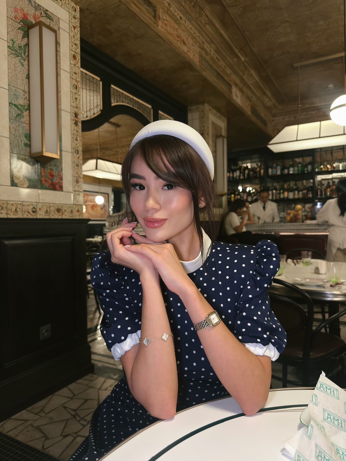 Aidana sits at a restaurant table, chin on clasped hands, wearing a white headband and a navy blue polka-dot dress with puffed sleeves.