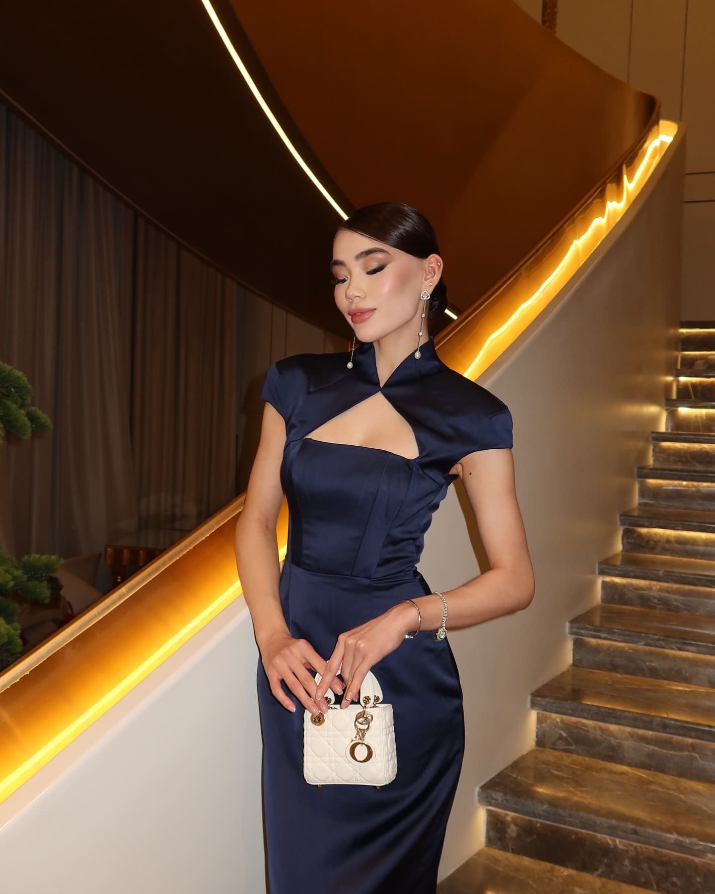 A woman with a sleek bun stands on a staircase in a navy blue satin dress with a high collar and keyhole cutout, holding a white quilted handbag.