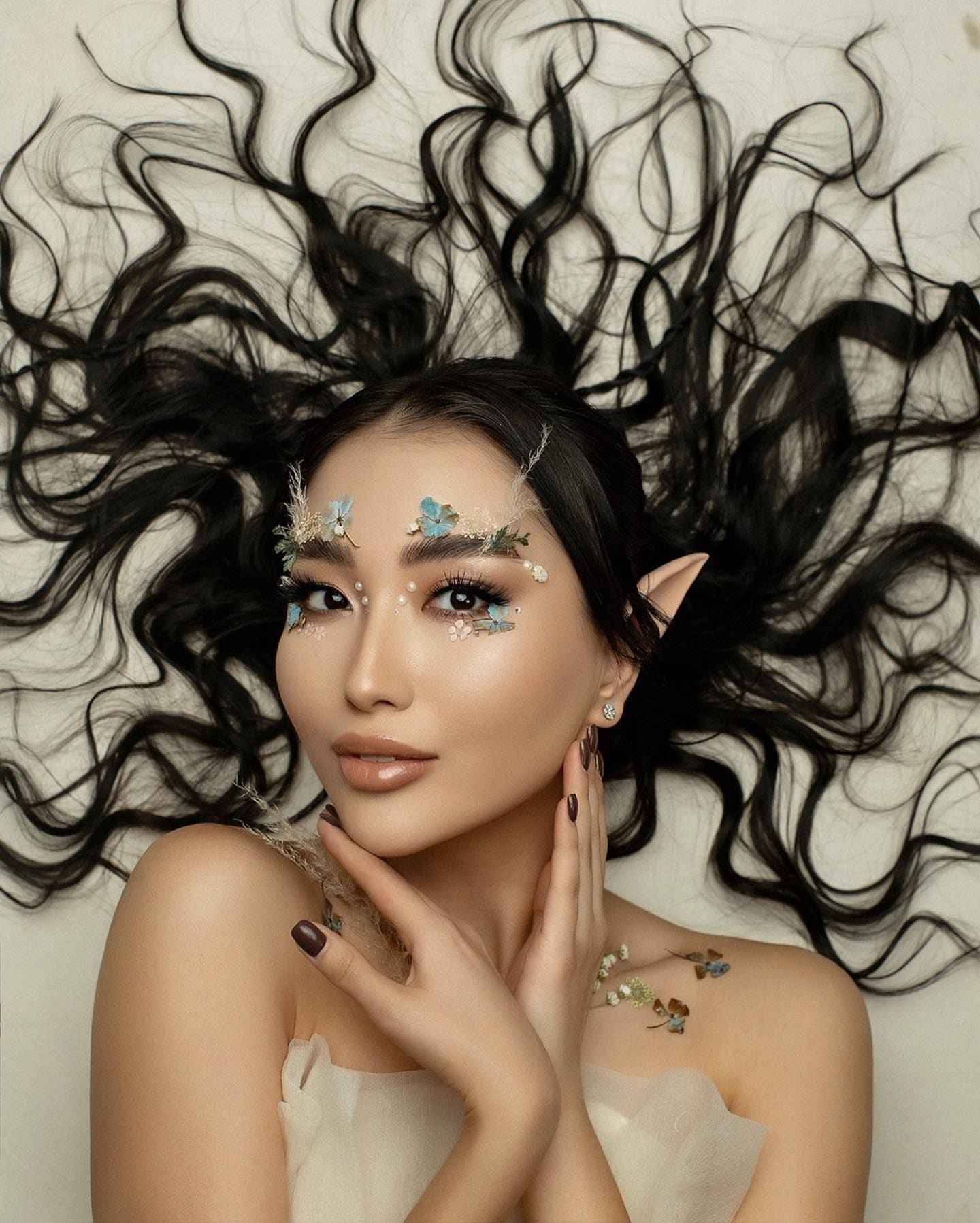 A woman with elf ears lies on a white surface in a strapless white garment with floral face decorations.