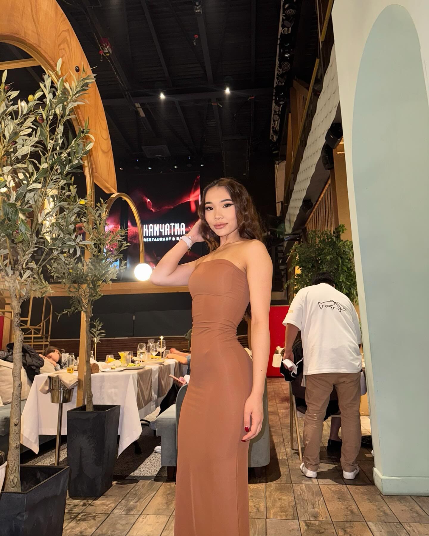 In a restaurant, she poses in a strapless floor-length brown dress, one hand raised near her neck, hair down and wavy.