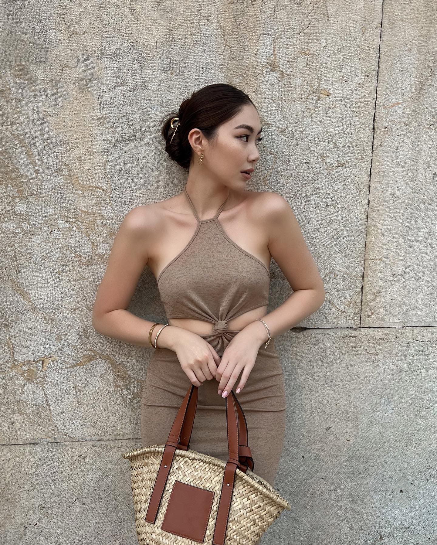 Woman in a beige halter crop top and skirt stands by a concrete wall, holding a straw tote bag.