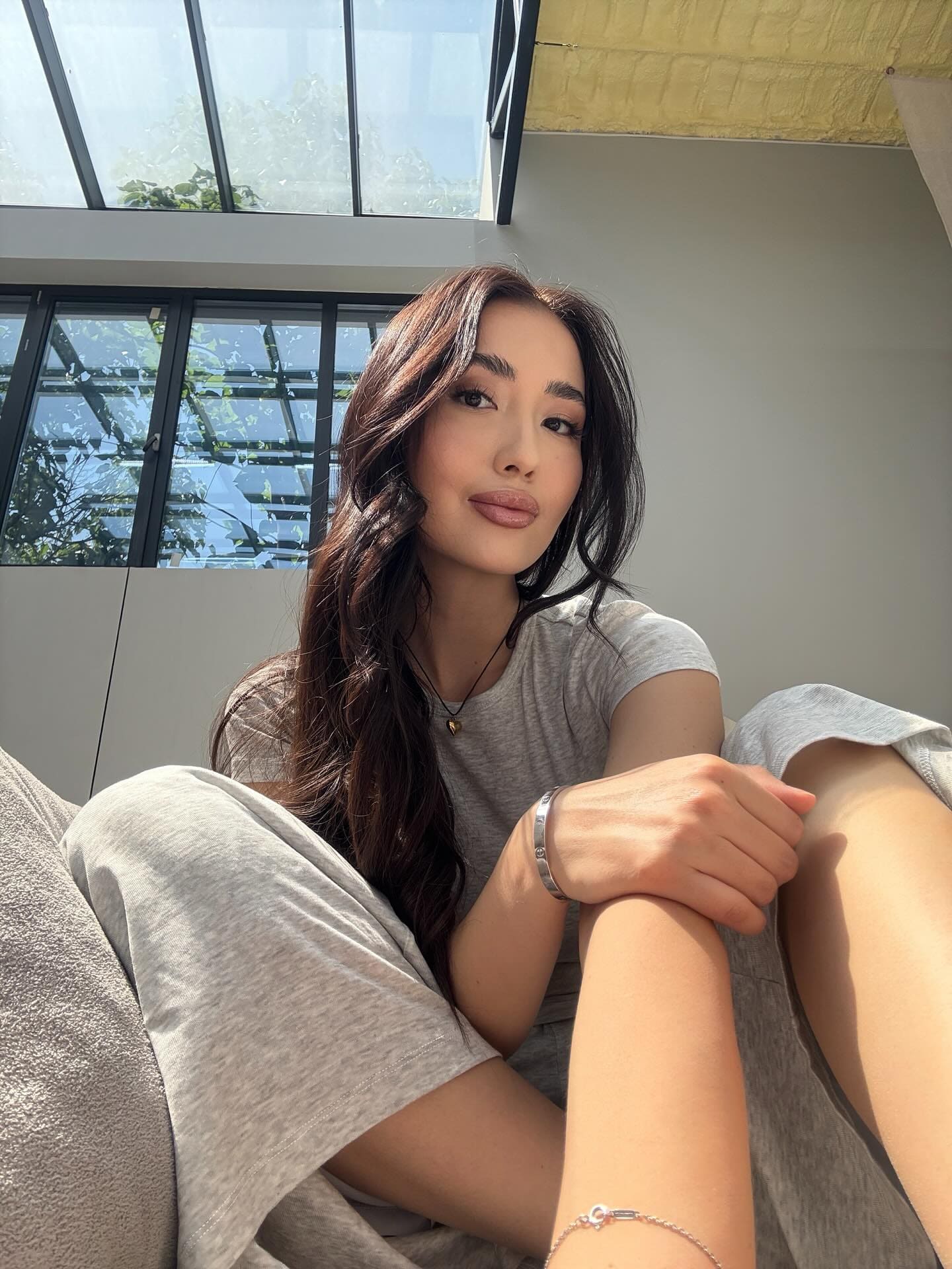 Erke sits indoors in a grey t-shirt and sweatpants, knees drawn up, in front of large windows.