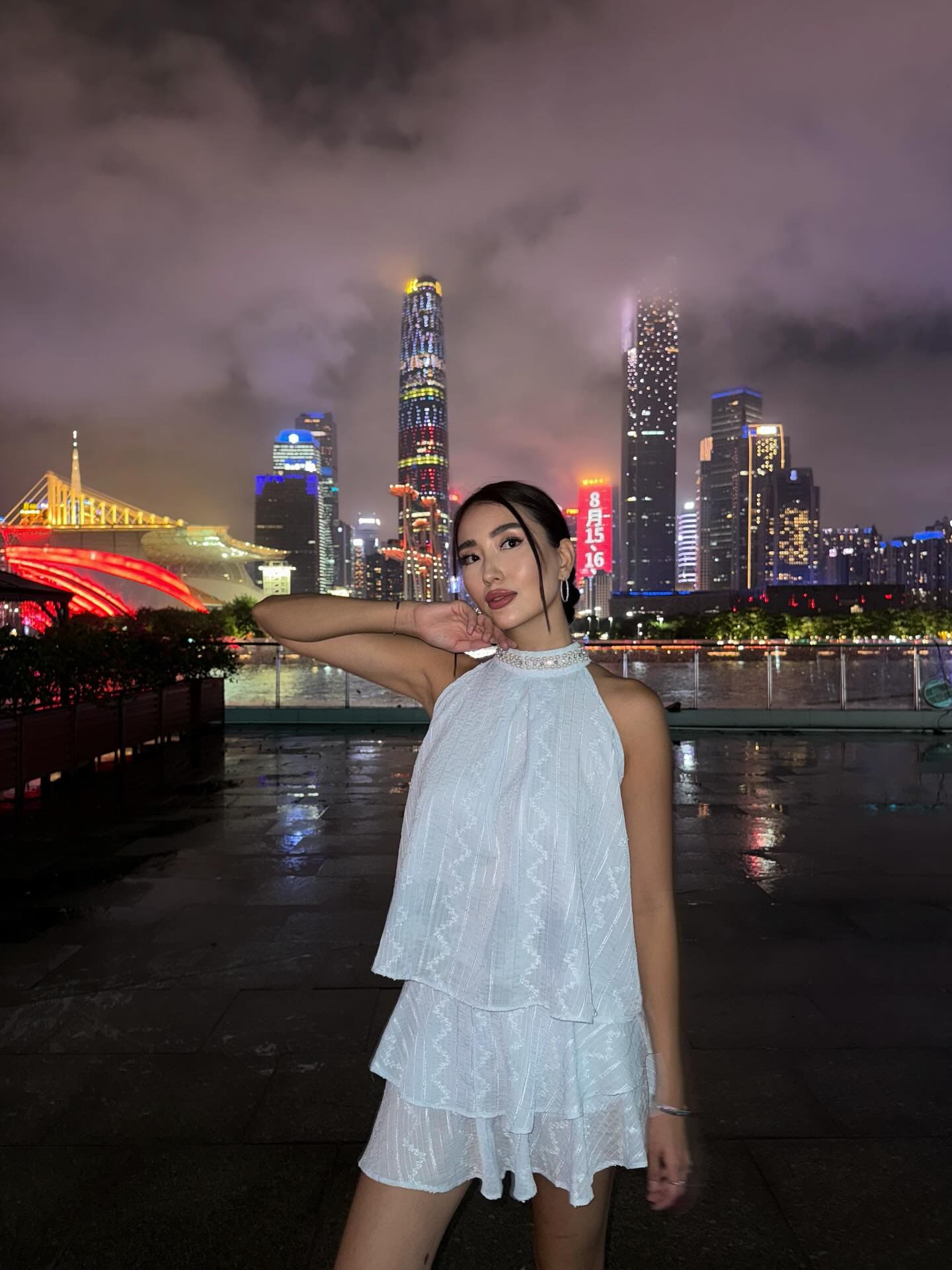 A woman in a white tiered dress stands outdoors at night against a city skyline with one hand raised.