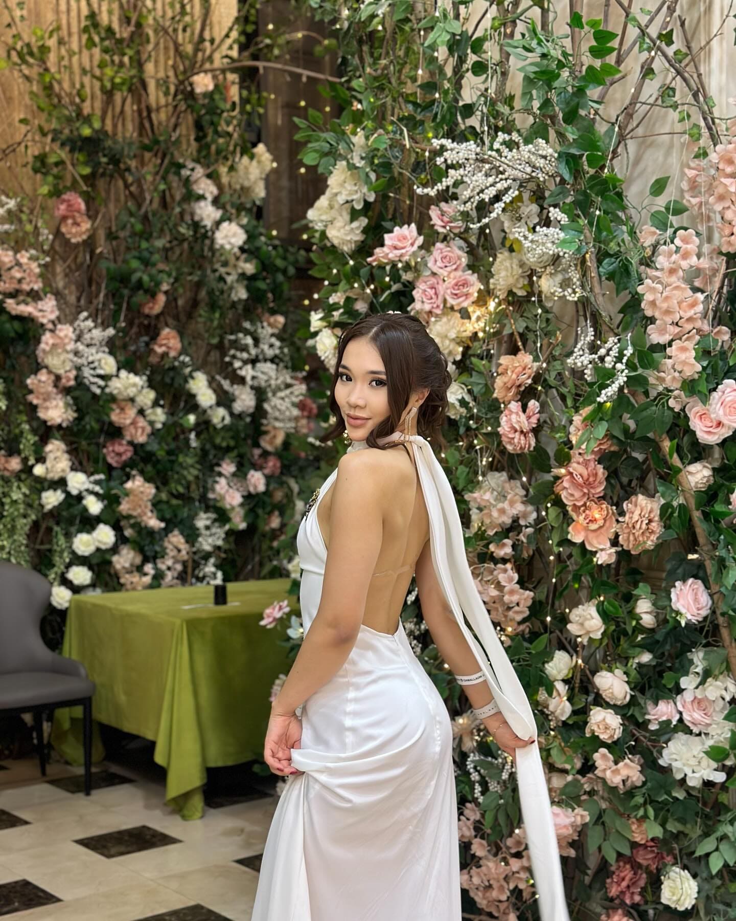 In profile against a flower wall, she wears a white satin halter gown with an open back, her hair styled in an updo.