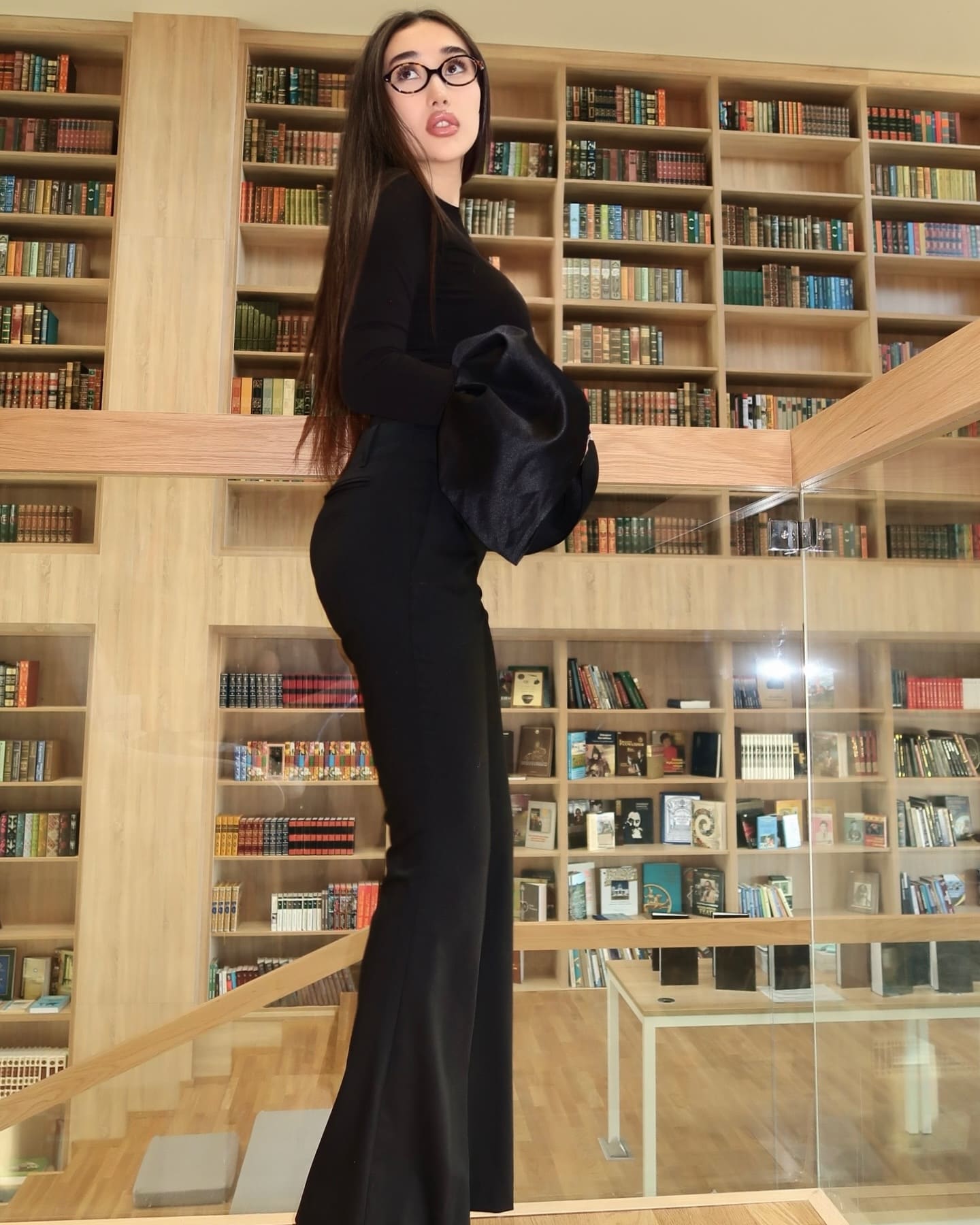 Wearing glasses and a black long-sleeved top with flared trousers, a woman stands in profile holding a jacket in front of wooden bookshelves.
