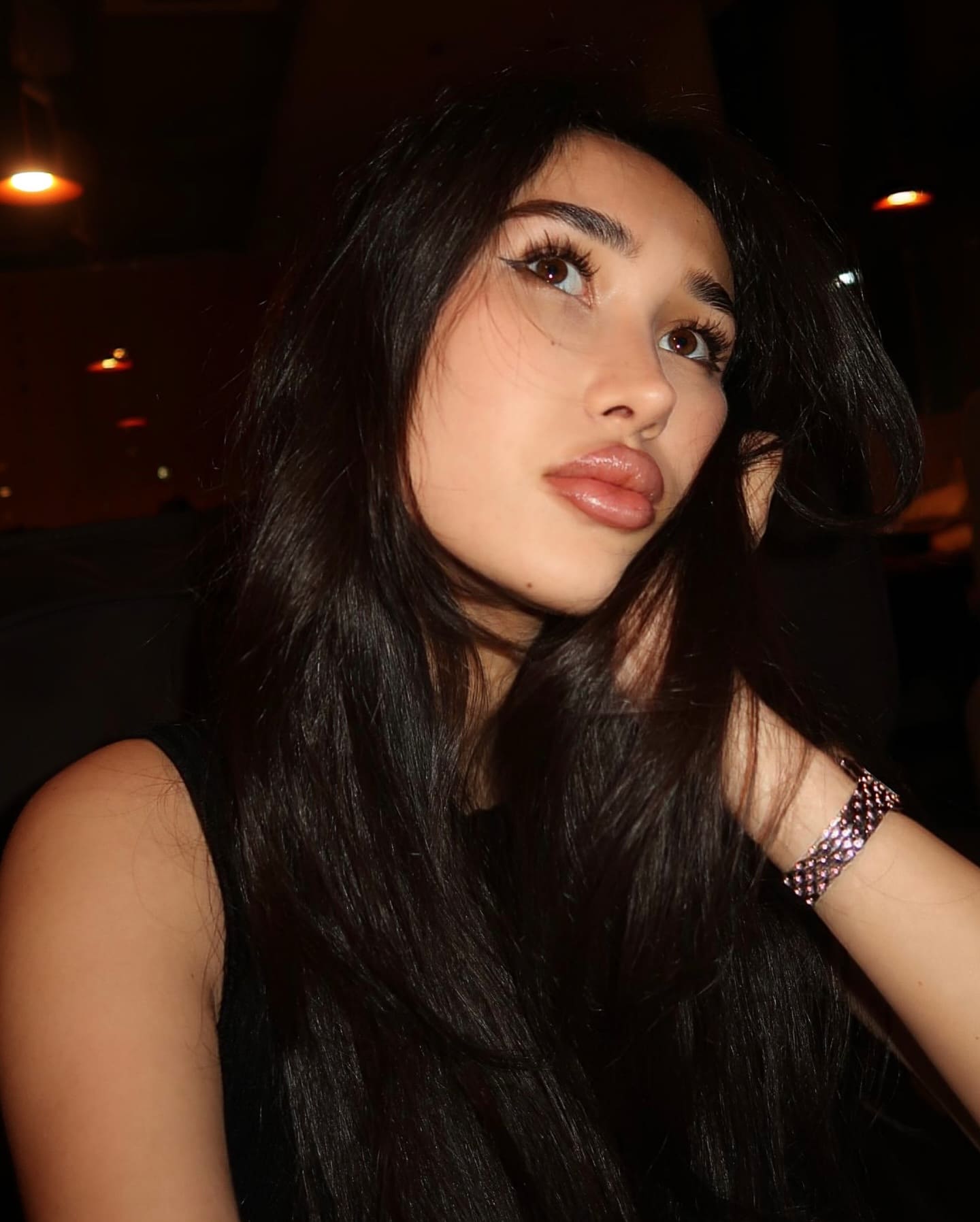 In a dimly lit setting, a woman in a black sleeveless top poses with one hand raised near her face, wearing a metallic bracelet.